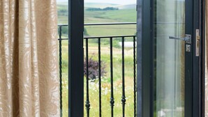 A balcony with a view of green fields, a black door, and beige curtains.