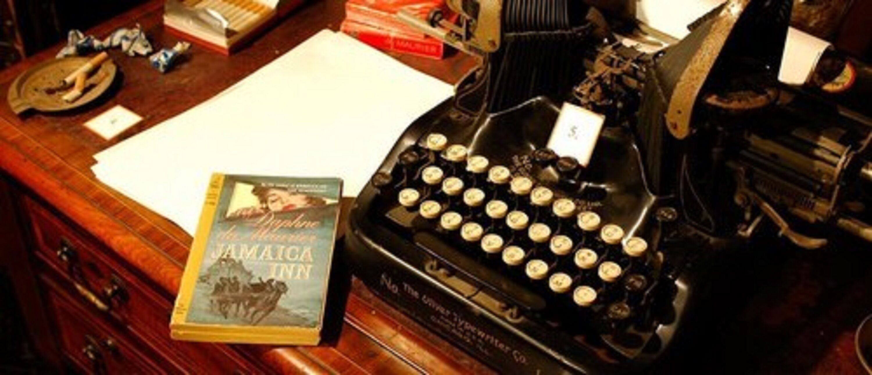 Une machine à écrire ancienne, un livre intitulé « Jamaica Inn », et un bureau en bois avec des papiers et une loupe.