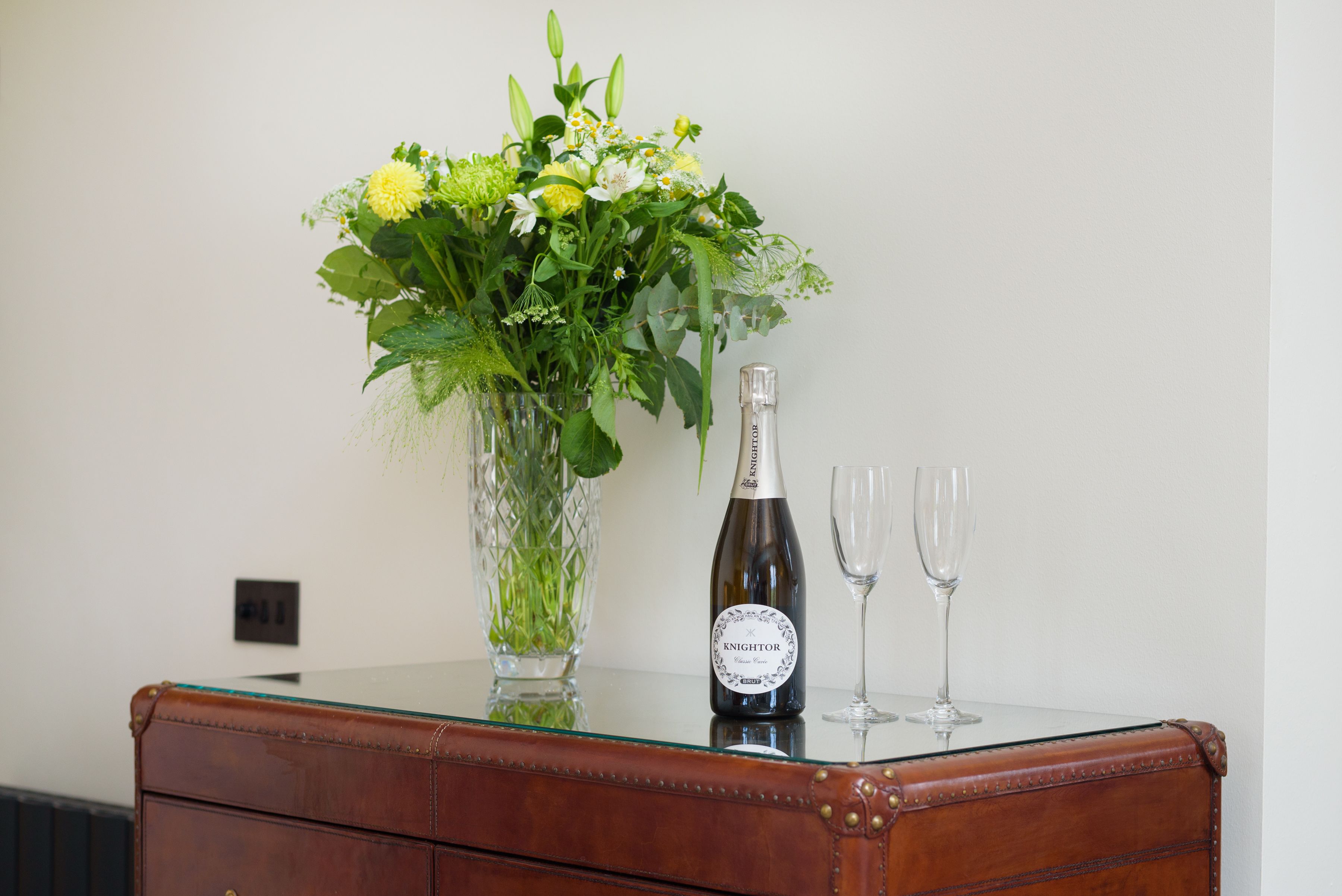 A wooden dresser with a glass top, a bottle of wine, two wine glasses, and a vase with flowers.