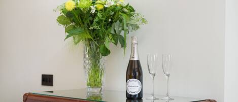 A wooden dresser with a glass top, a bottle of wine, two wine glasses, and a vase with flowers.