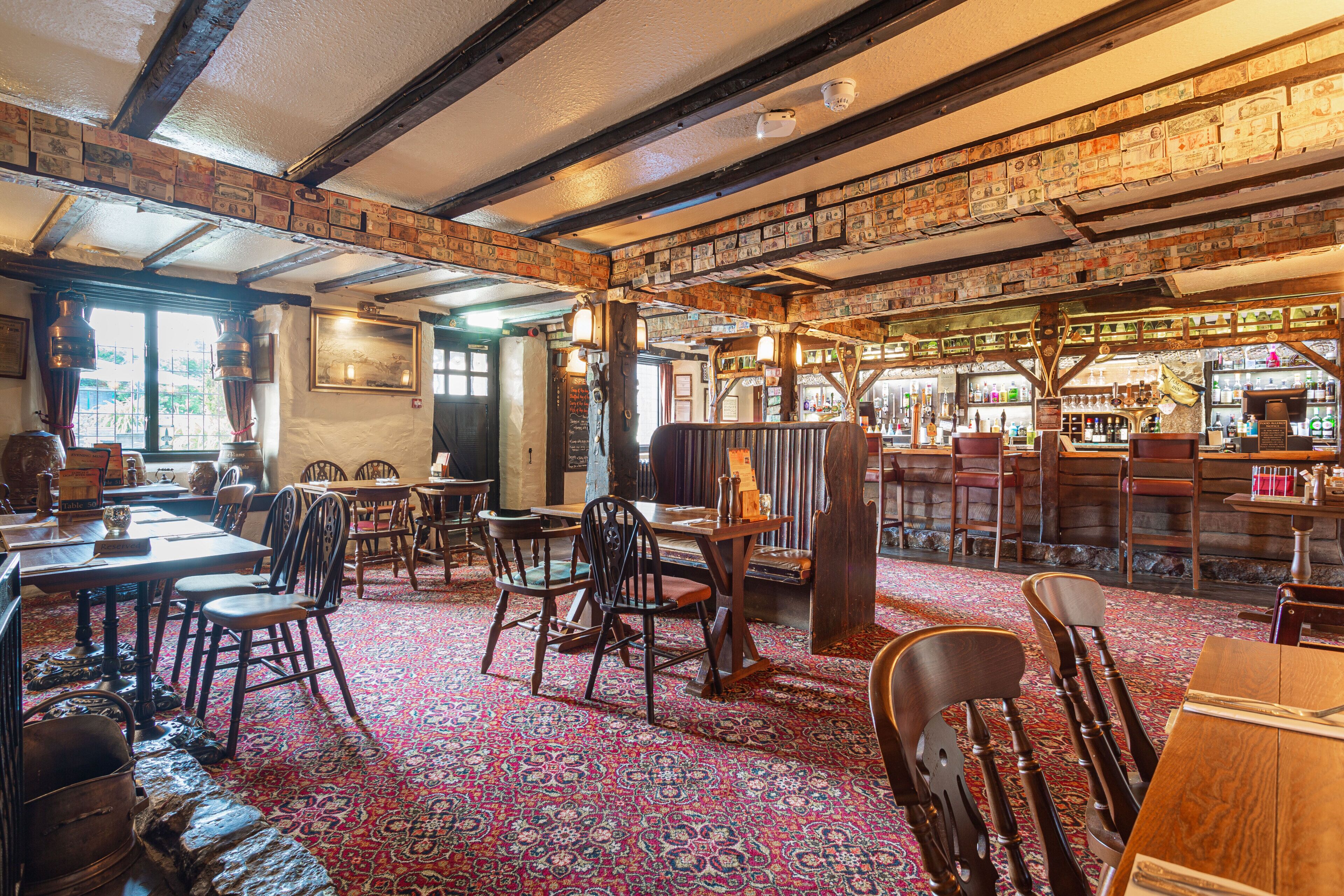 Interior de un pub tradicional con vigas de madera, alfombra estampada y una zona de bar con botellas.