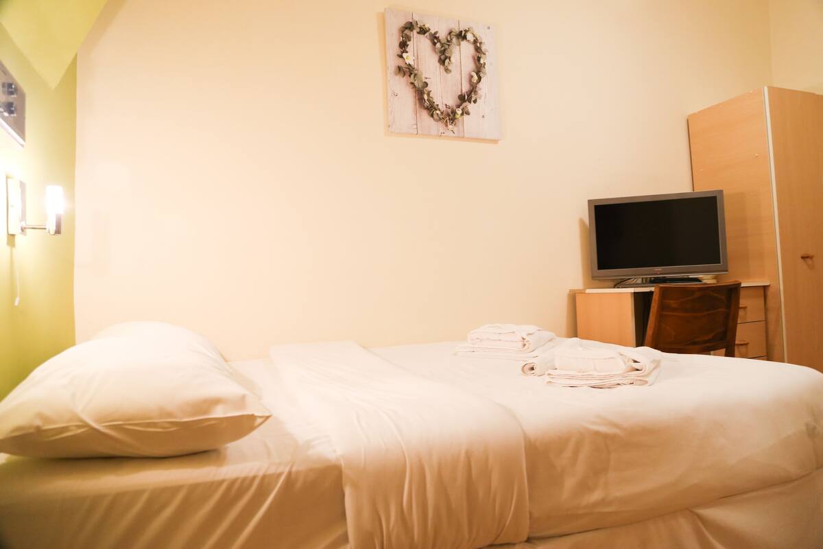 A neatly made bed with white linens, a heart-shaped decoration on the wall, and a television on a small table beside the bed.