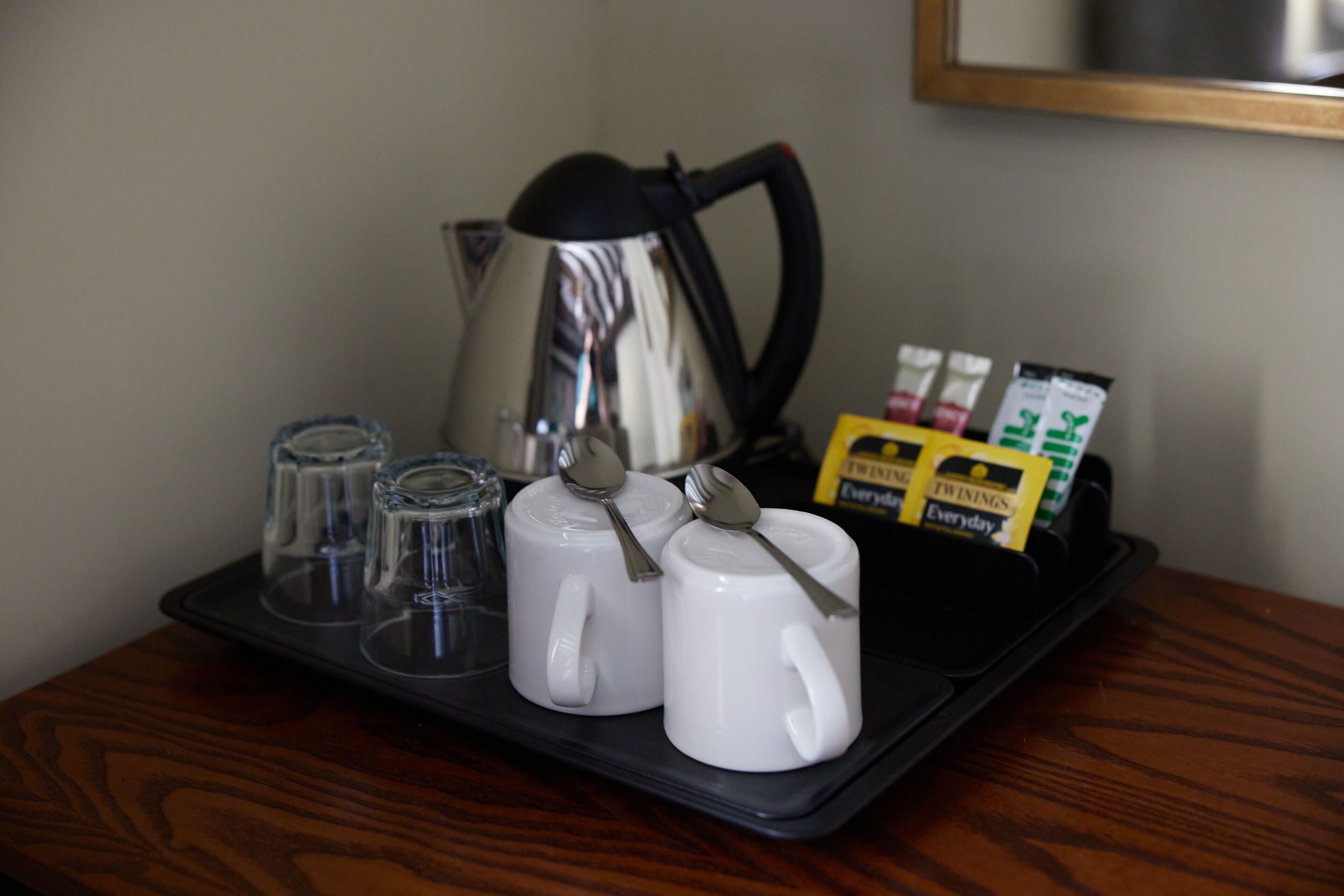 A tray with two white mugs, a glass, a kettle, and some packets on a wooden table.