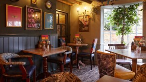 Ein gemütliches Restaurantambiente mit Holztischen und -stühlen, gerahmten Bildern an den Wänden und einem großen Fenster mit Blick auf Grünflächen.
