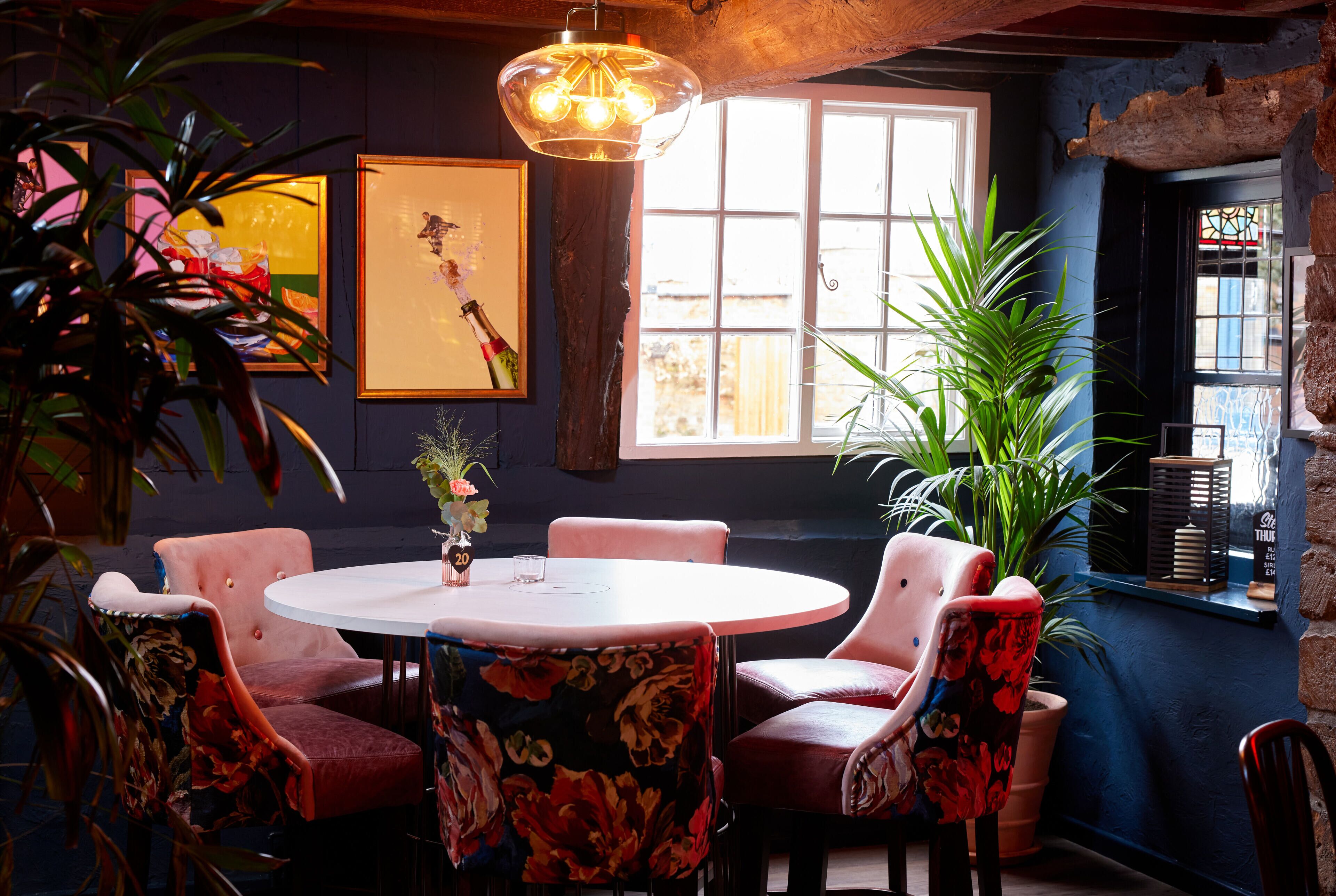A cozy dining area with a round table and floral chairs, surrounded by potted plants and framed artwork on the walls.