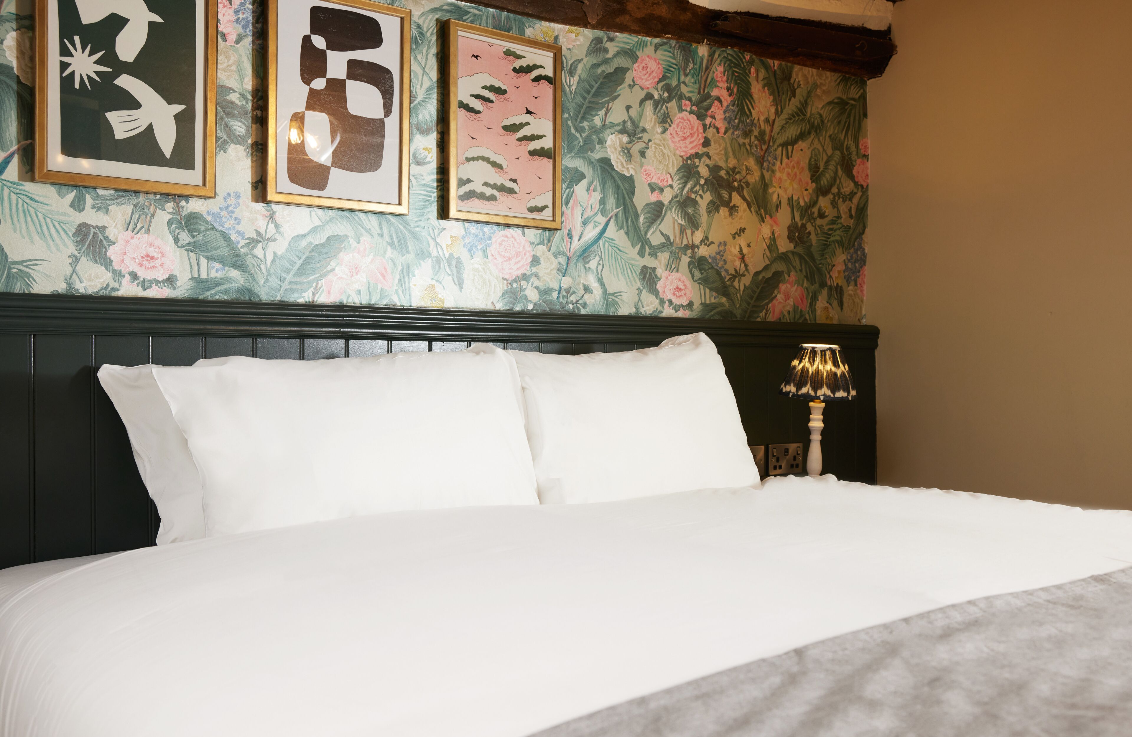 A neatly made bed with white linens in a room with floral wallpaper and framed artwork on the wall.
