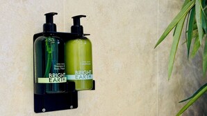 Due flaconi di shampoo e balsamo Bright Earth su un portaoggetti nero fissato a parete in un bagno con piastrelle color crema.