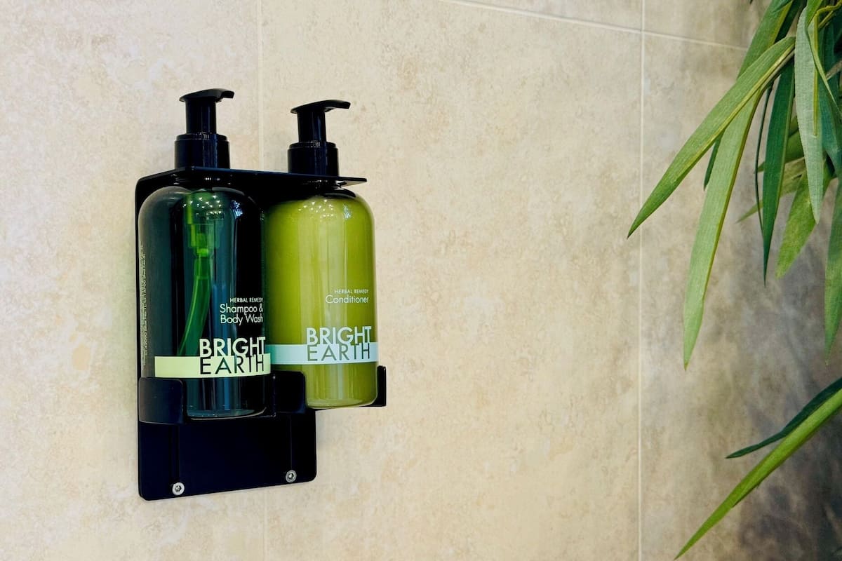 Two bottles of Bright Earth shampoo and conditioner on a black wall-mounted holder in a bathroom with beige tiles.