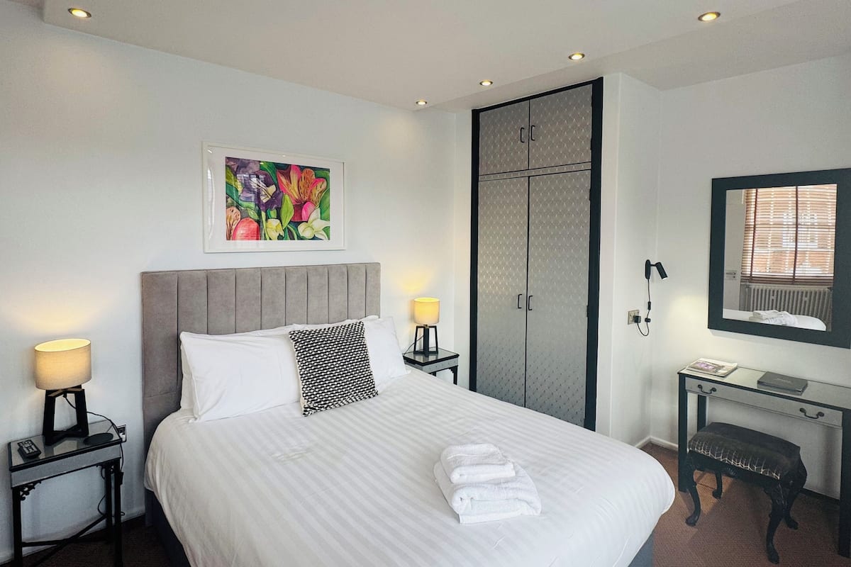 A hotel room with a large bed, a nightstand with a lamp, a desk with a chair, a wardrobe, and a framed picture on the wall.