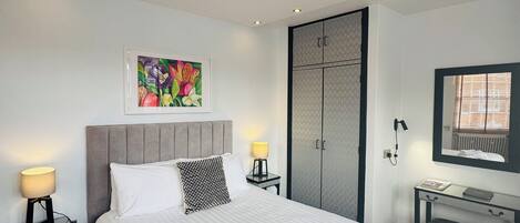 Een hotelkamer met een groot bed, een nachtkastje met een lamp, een bureau met een stoel, een kledingkast en een ingelijste foto aan de muur.