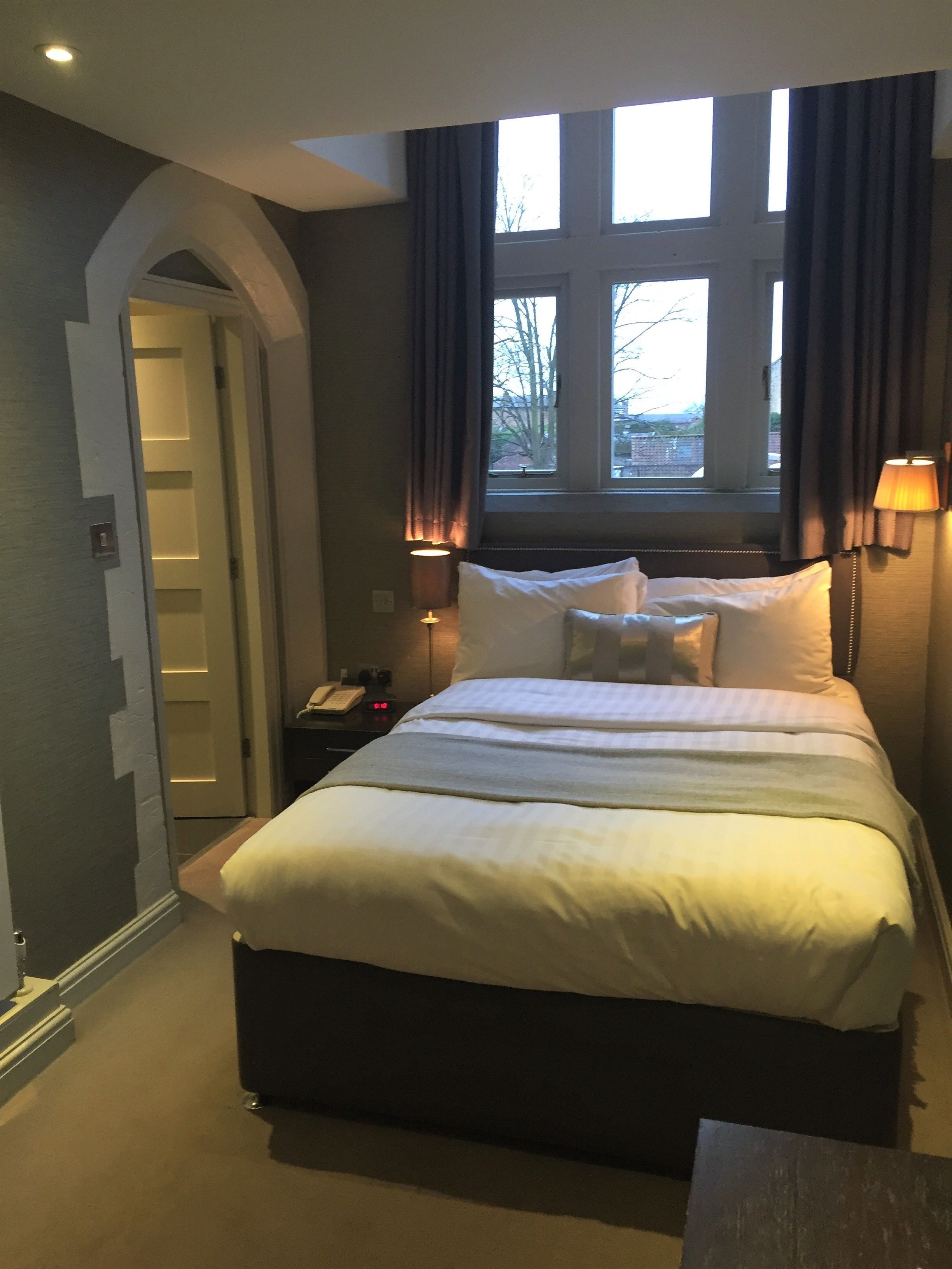 A hotel room with a large bed, a nightstand with a lamp, a window with curtains, and a door leading to another room.