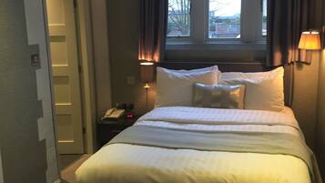 A hotel room with a large bed, a nightstand with a lamp, a window with curtains, and a door leading to another room.