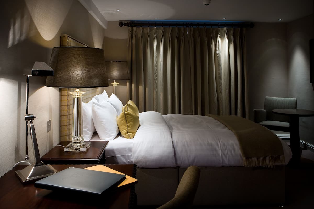 A hotel room with a bed, bedside tables, a desk, a chair, and a large window with curtains.