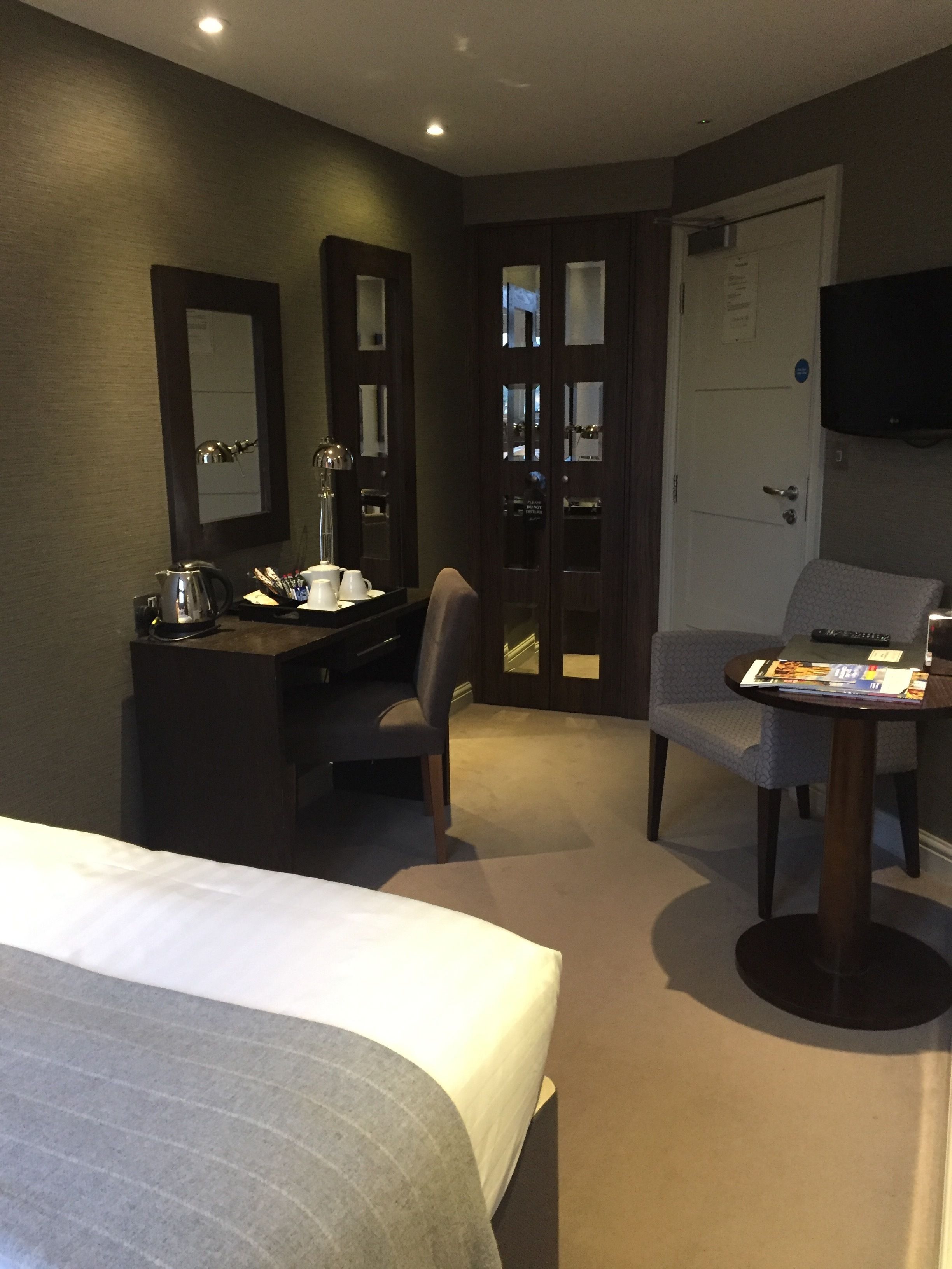 Habitación de hotel con cama, un escritorio con silla, una mesita con otra silla, televisión y una puerta con espejo.