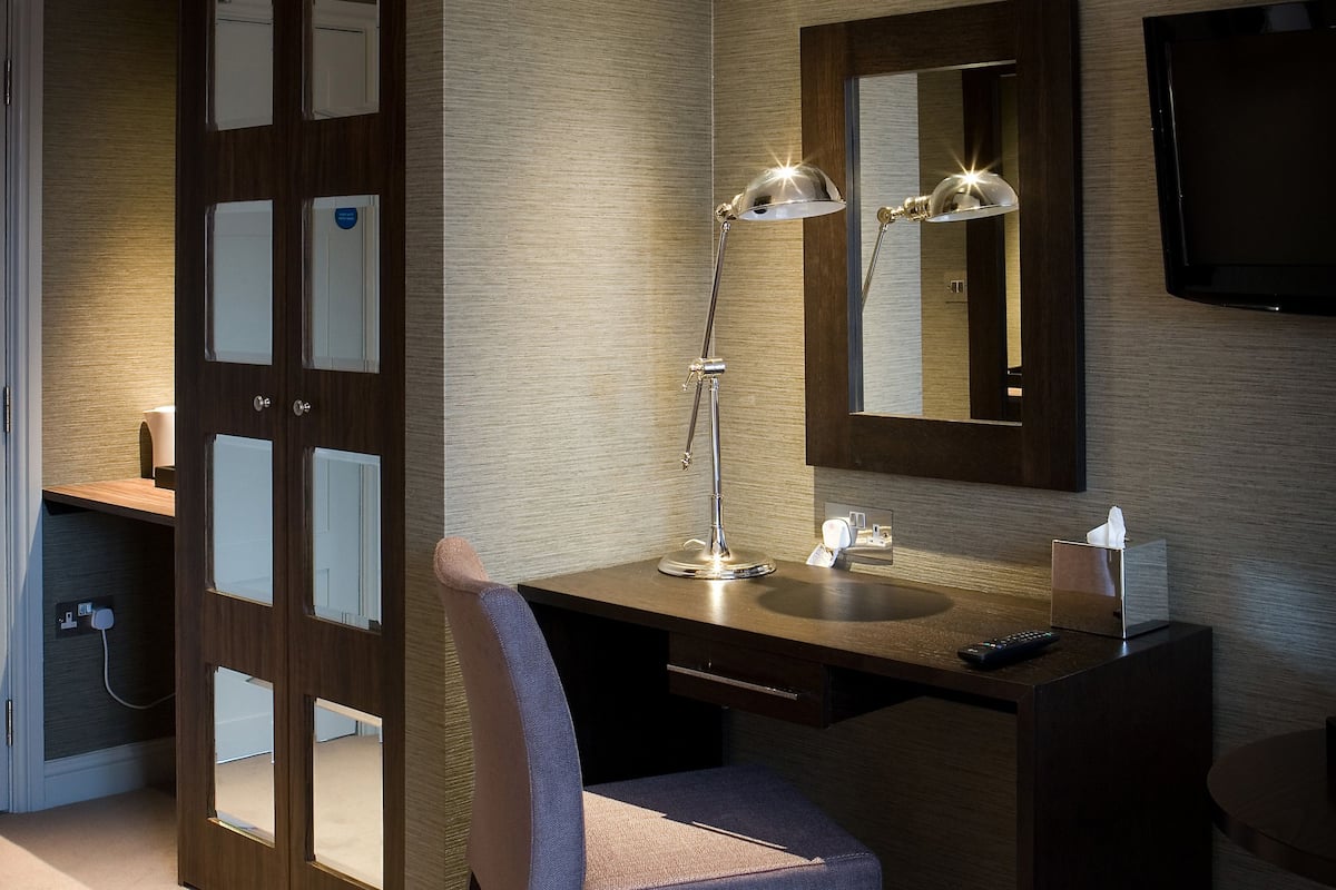 A hotel room with a desk, chair, lamp, mirror, TV, and a door with glass panels.