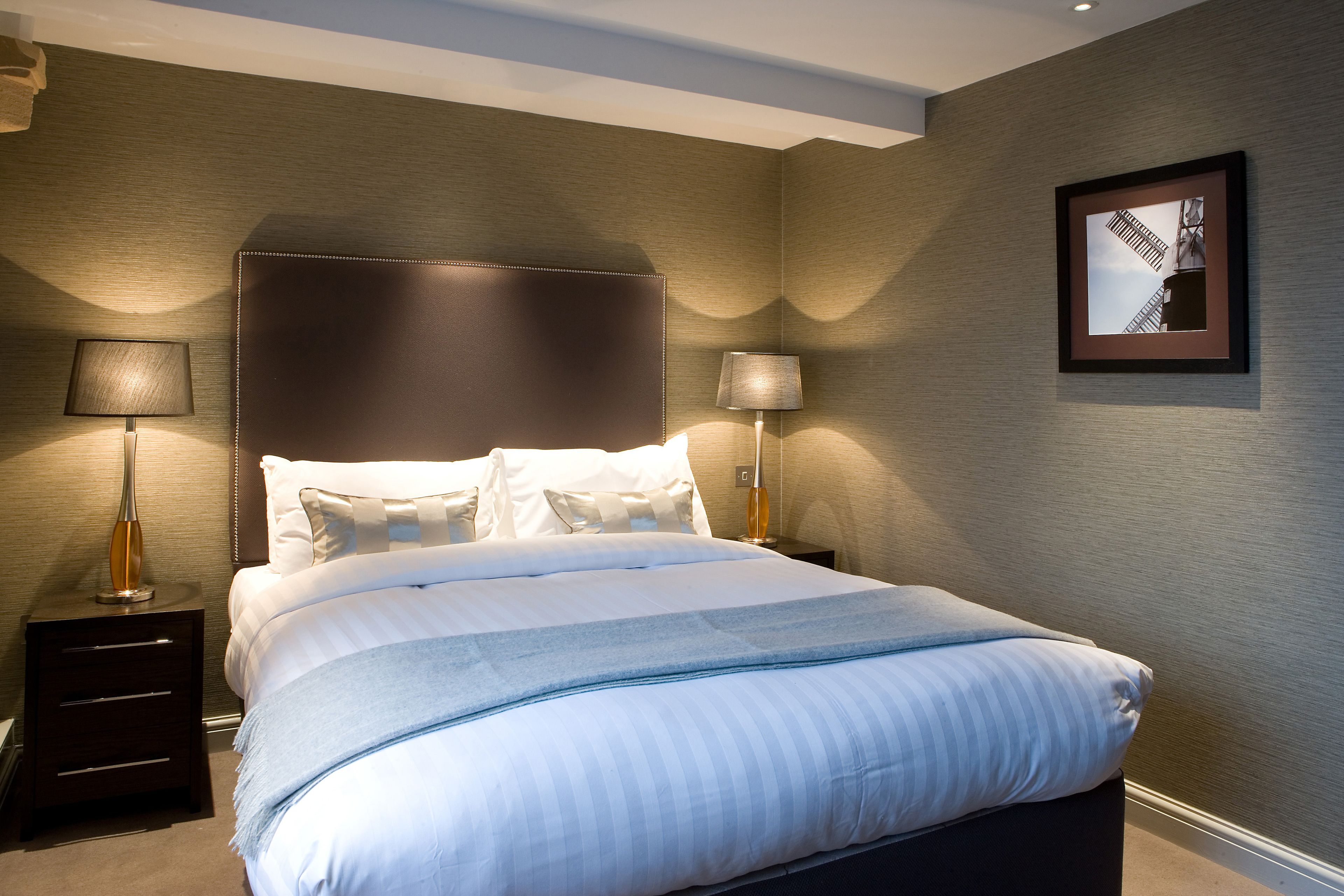 A neatly made bed with white linens and a blue throw blanket, flanked by two bedside tables with lamps, in a room with a textured wall and a framed picture.