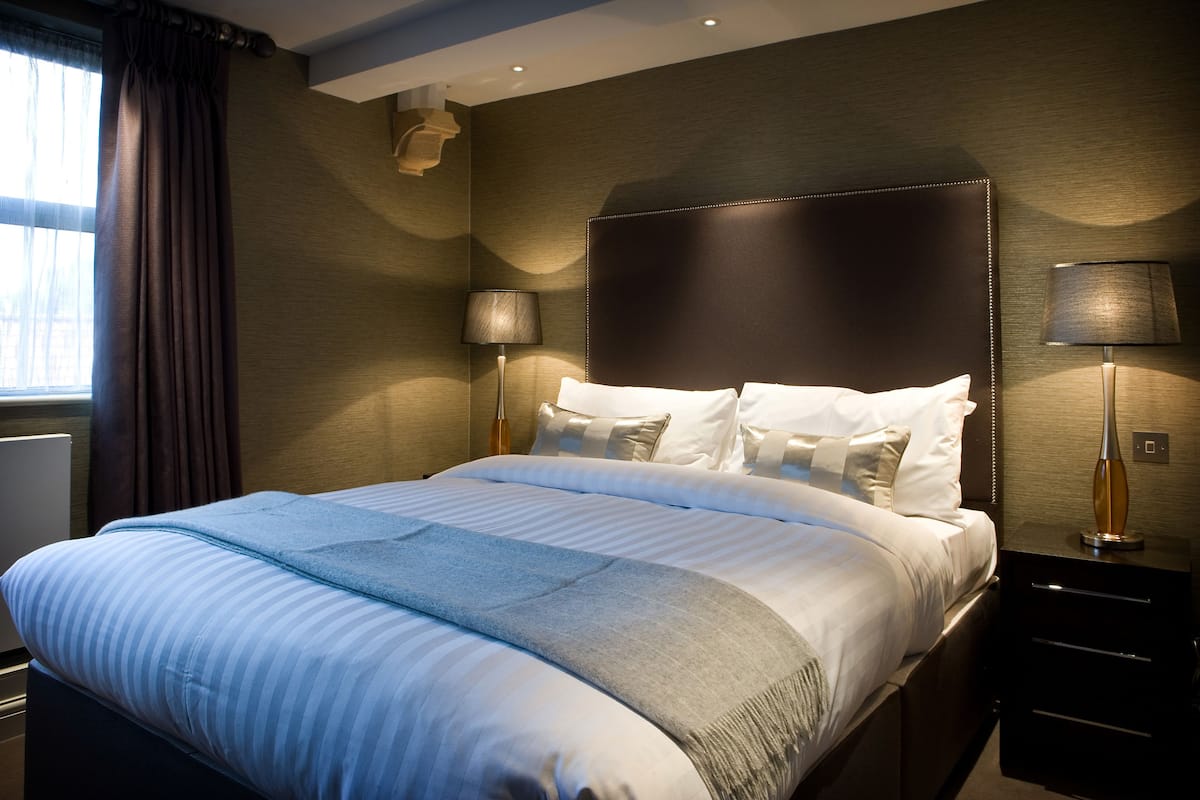 A neatly made bed with a headboard, two bedside lamps, and a window with curtains.