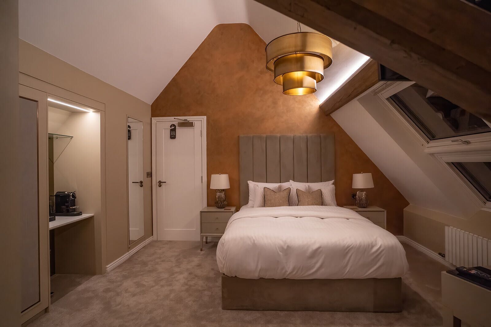 A bedroom with a sloped ceiling, a bed with white bedding, two bedside tables with lamps, and a small shelf with a coffee maker.