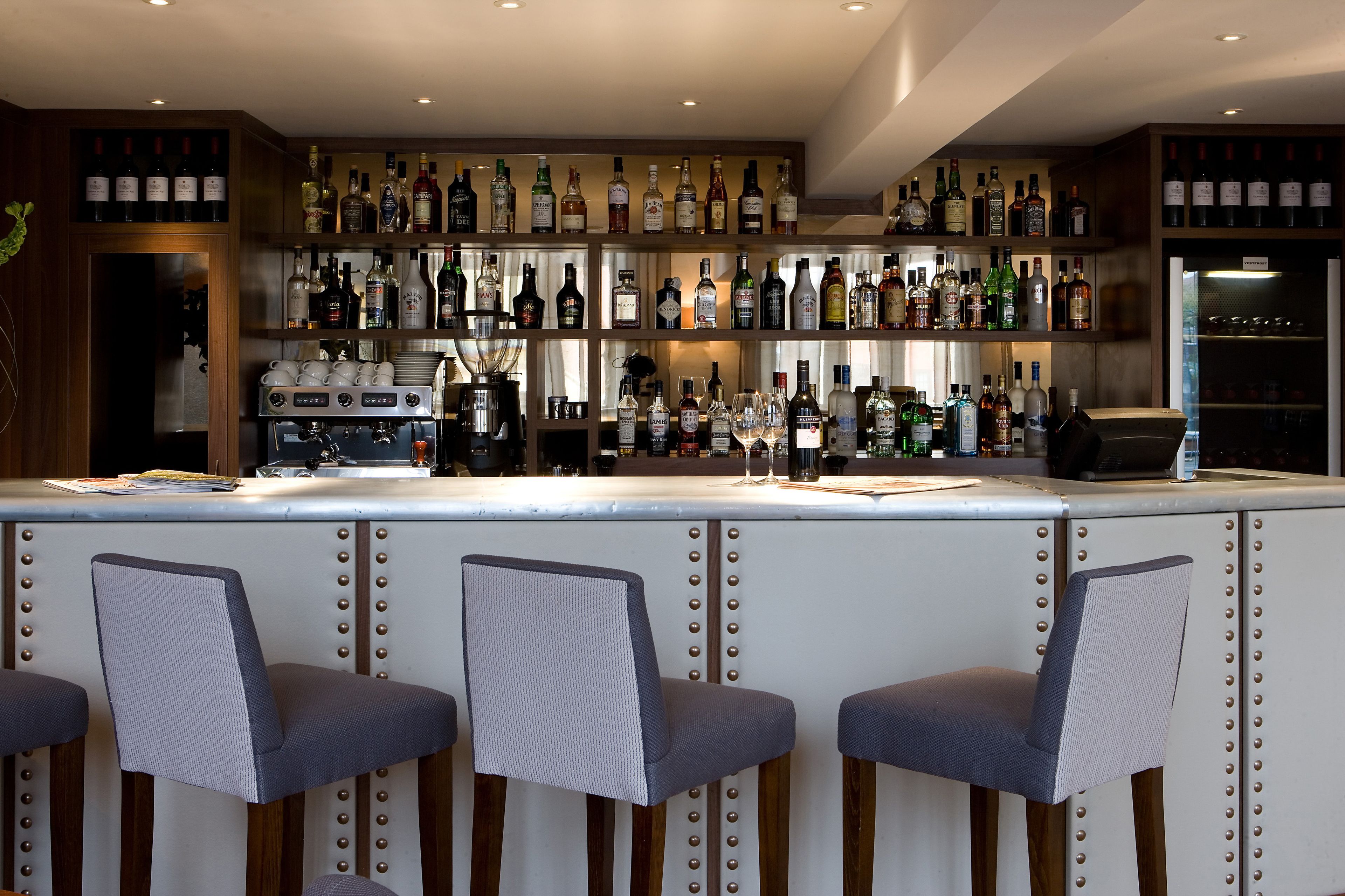 Una zona de bar con varias estanterías llenas de botellas de licores, una máquina de café y tres taburetes tapizados.