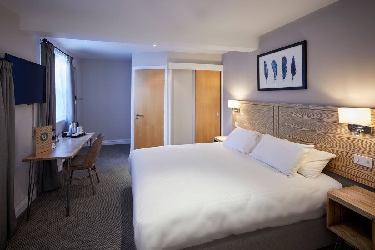 A hotel room with a large bed, a desk, a chair, a TV, and a framed artwork on the wall.