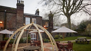 An outdoor patio area with wooden tables and chairs, surrounded by brick walls and a large tree. - Childwall Abbey, by Marstons Inns  (Liverpool)