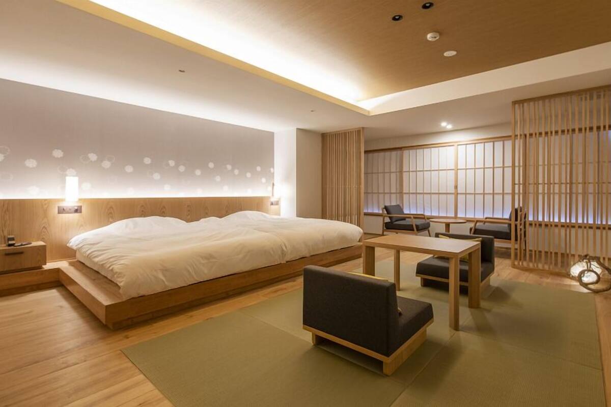 A modern hotel room with a large bed, a small desk, a chair, and a view of a traditional Japanese room.