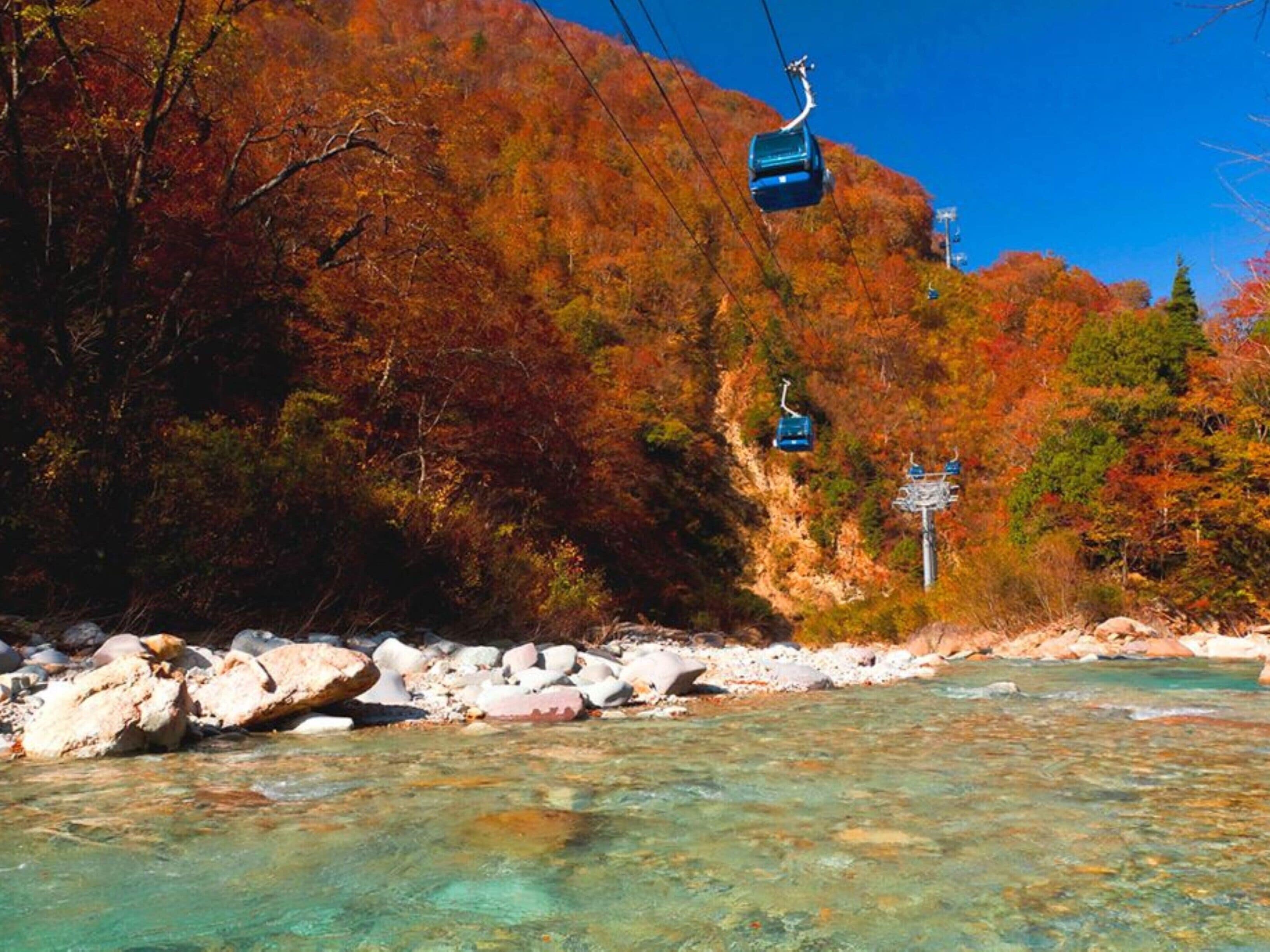 Ein klarer Fluss mit Steinen, ein Berg mit Herbstlaub und eine Seilbahn.