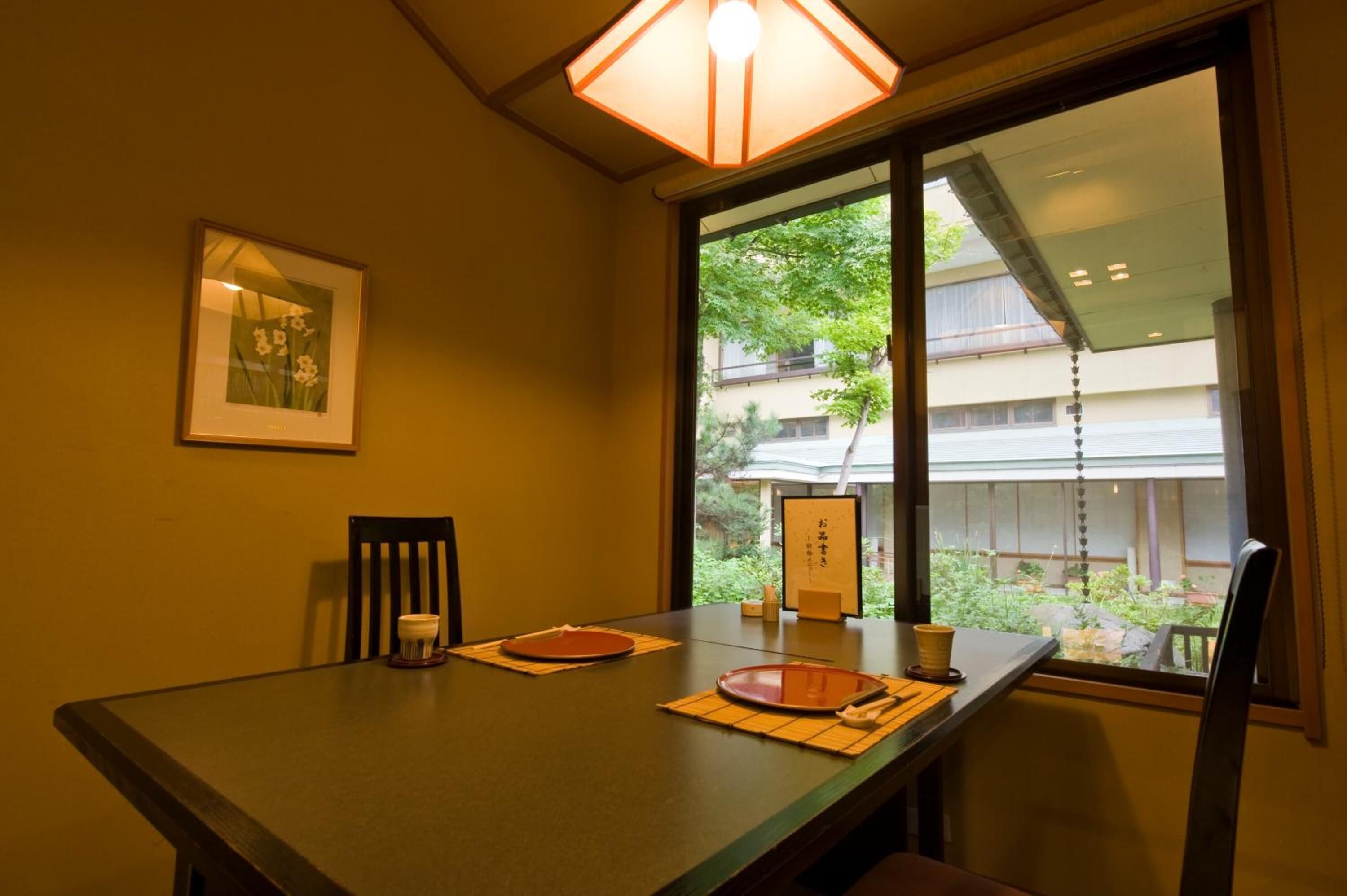 Een eettafel met een placemat, een inlijsting met een foto aan de muur, en een raam met uitzicht op groen en gebouwen.
