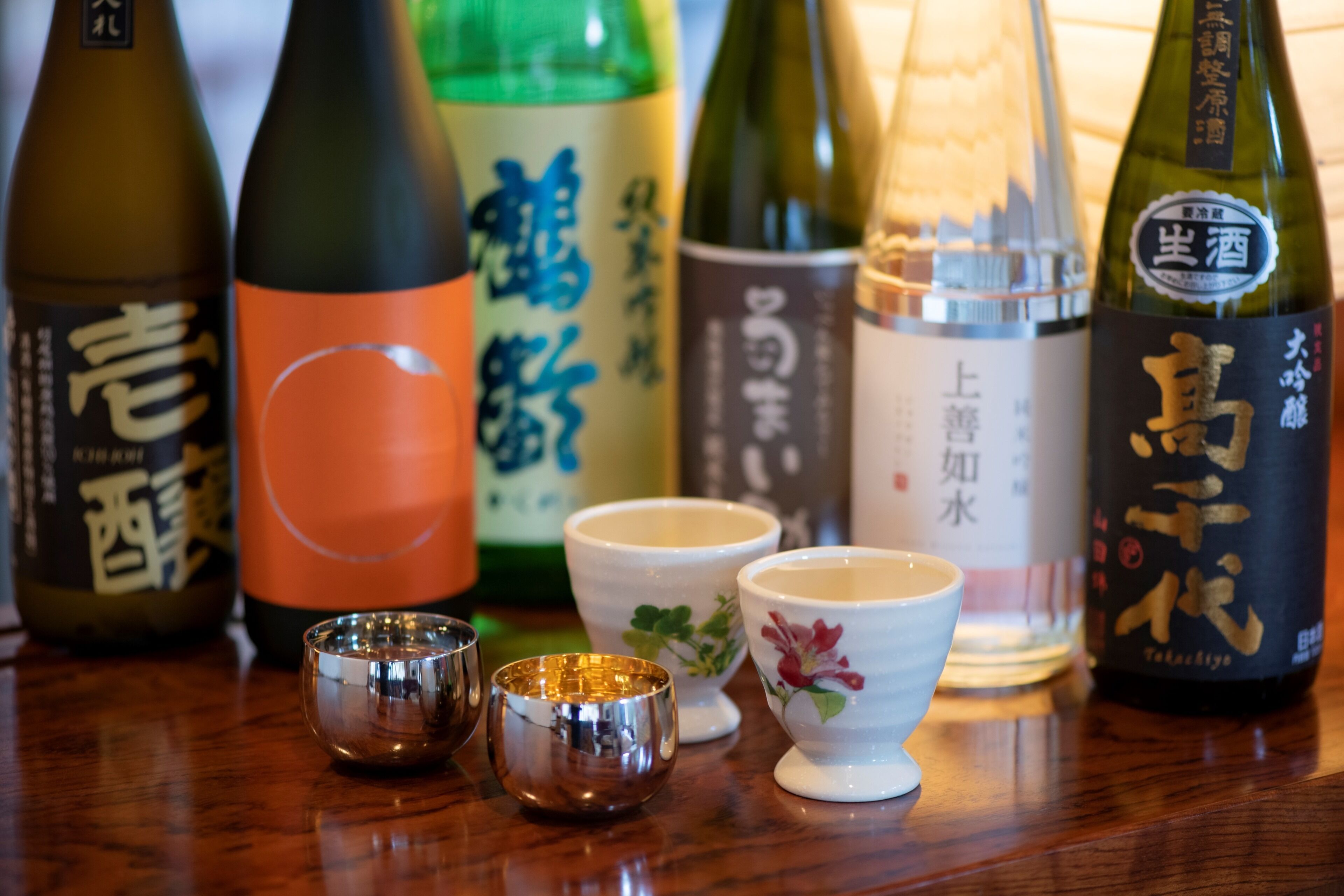 Variedad de botellas de sake y tazas tradicionales de té japonesas sobre una mesa de madera.