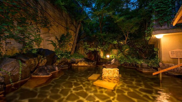 Un espace paisible avec des bains chauds naturels, comprenant un banc en bois, des rochers et une petite structure illuminée.