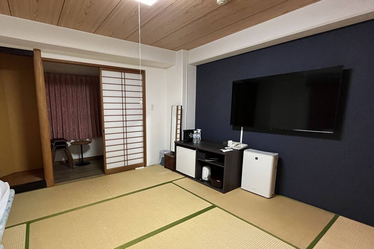 A hotel room with tatami flooring, a bed, a desk with a chair, a TV, and a sliding door.