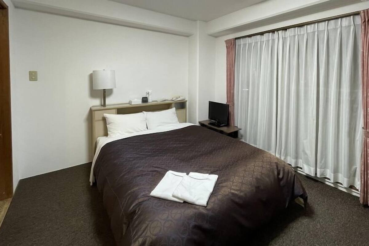 A hotel room with a bed, a bedside table with a lamp, a TV, and a window with curtains.