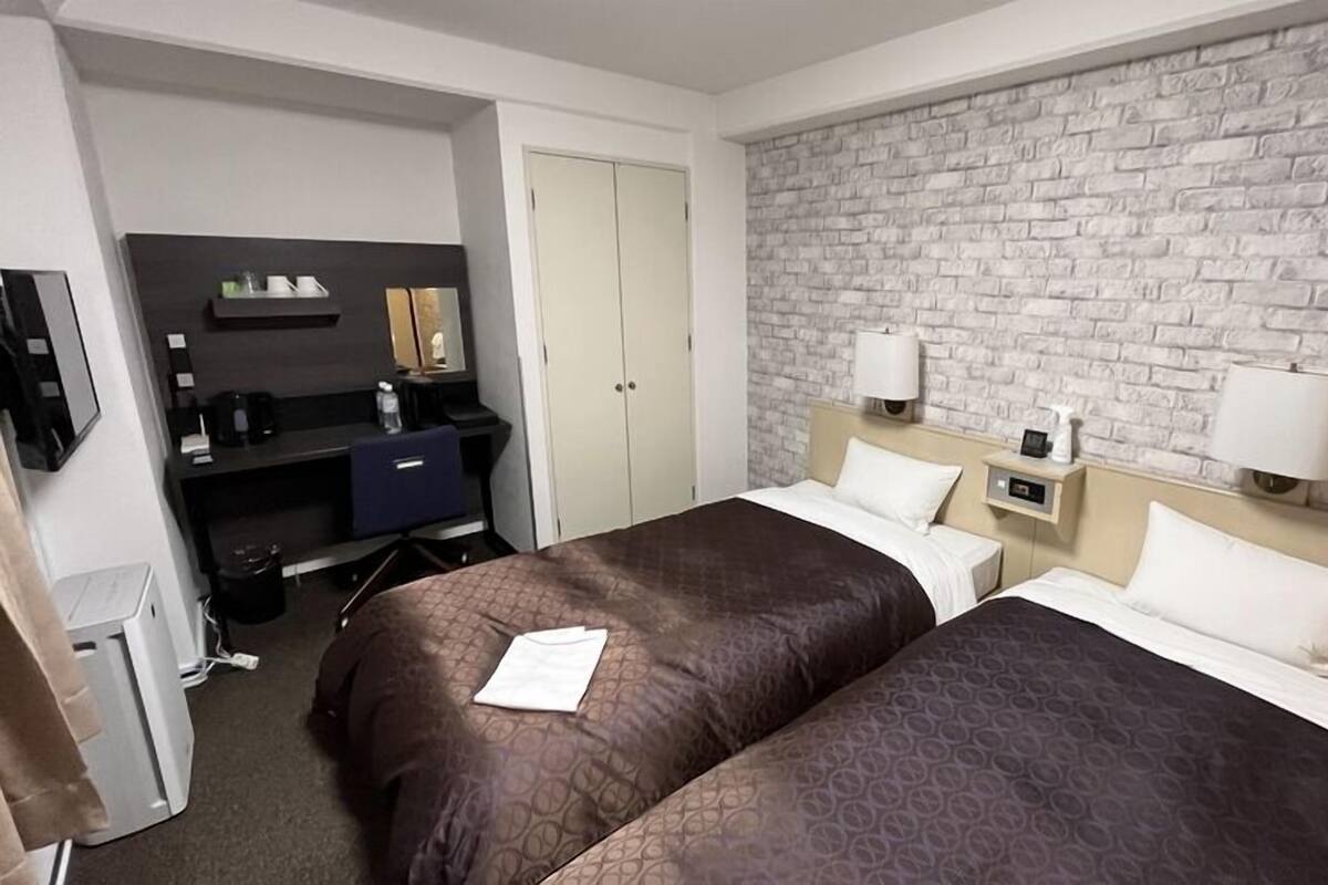 A hotel room with two beds, a desk, a mirror, a wardrobe, and a brick-patterned wall.