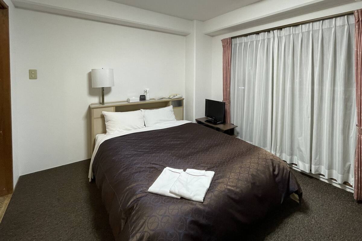 A hotel room with a bed, a bedside table with a lamp, a TV, and a window with curtains.