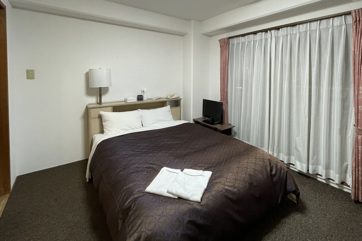 Quarto de hotel com cama, mesa de cabeceira com abajur, televisão e janela com cortinas.
