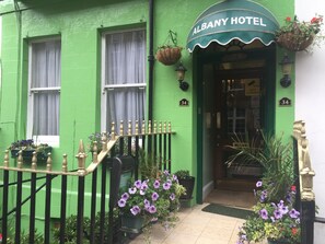 A green building with a sign reading "ALBANY HOTEL" above the entrance. There are potted plants and a small staircase leading to the door.