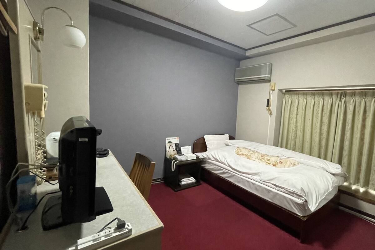 A hotel room with a bed, a desk with a computer, a telephone, and an air conditioning unit.