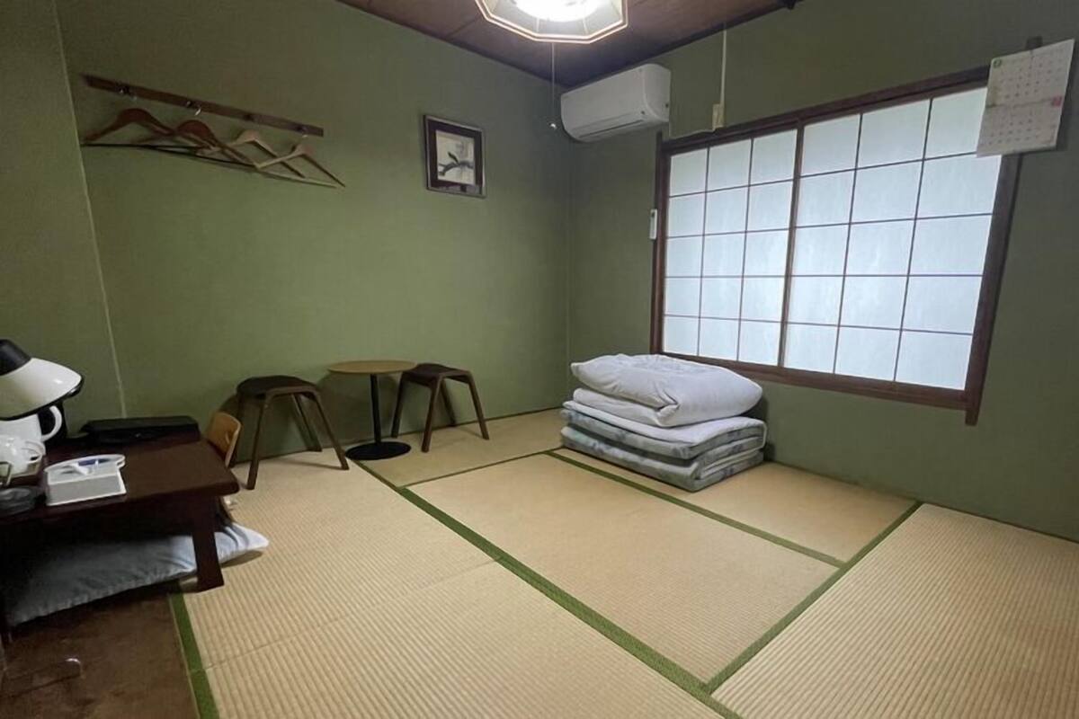 A room with tatami mats, a futon, a desk with a lamp, and a window with a view.