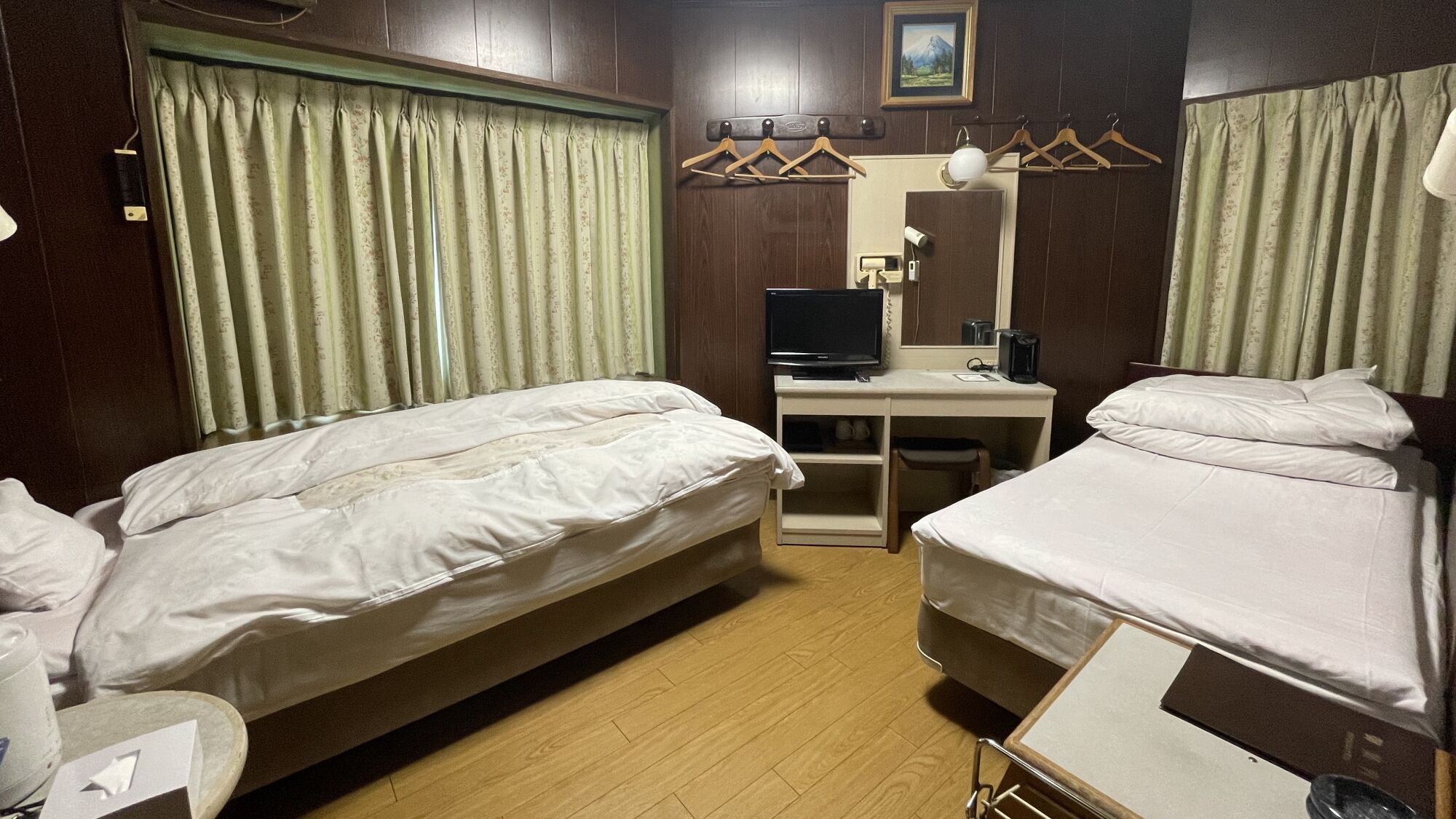 Ein Hotelzimmer mit zwei Betten, einem Schreibtisch mit Computer, einem Nachttisch und einem Fenster mit Vorhängen.