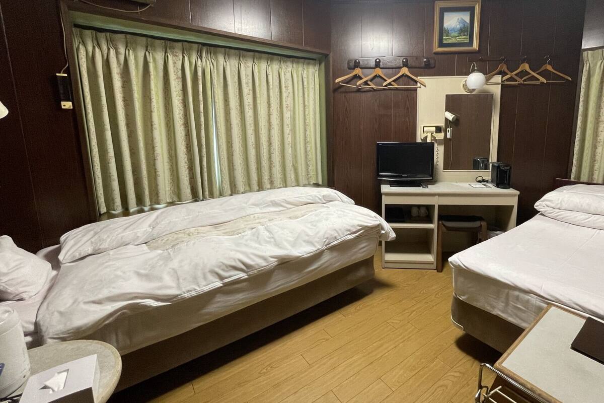 A hotel room with two beds, a desk with a computer, a bedside table, and a window with curtains.