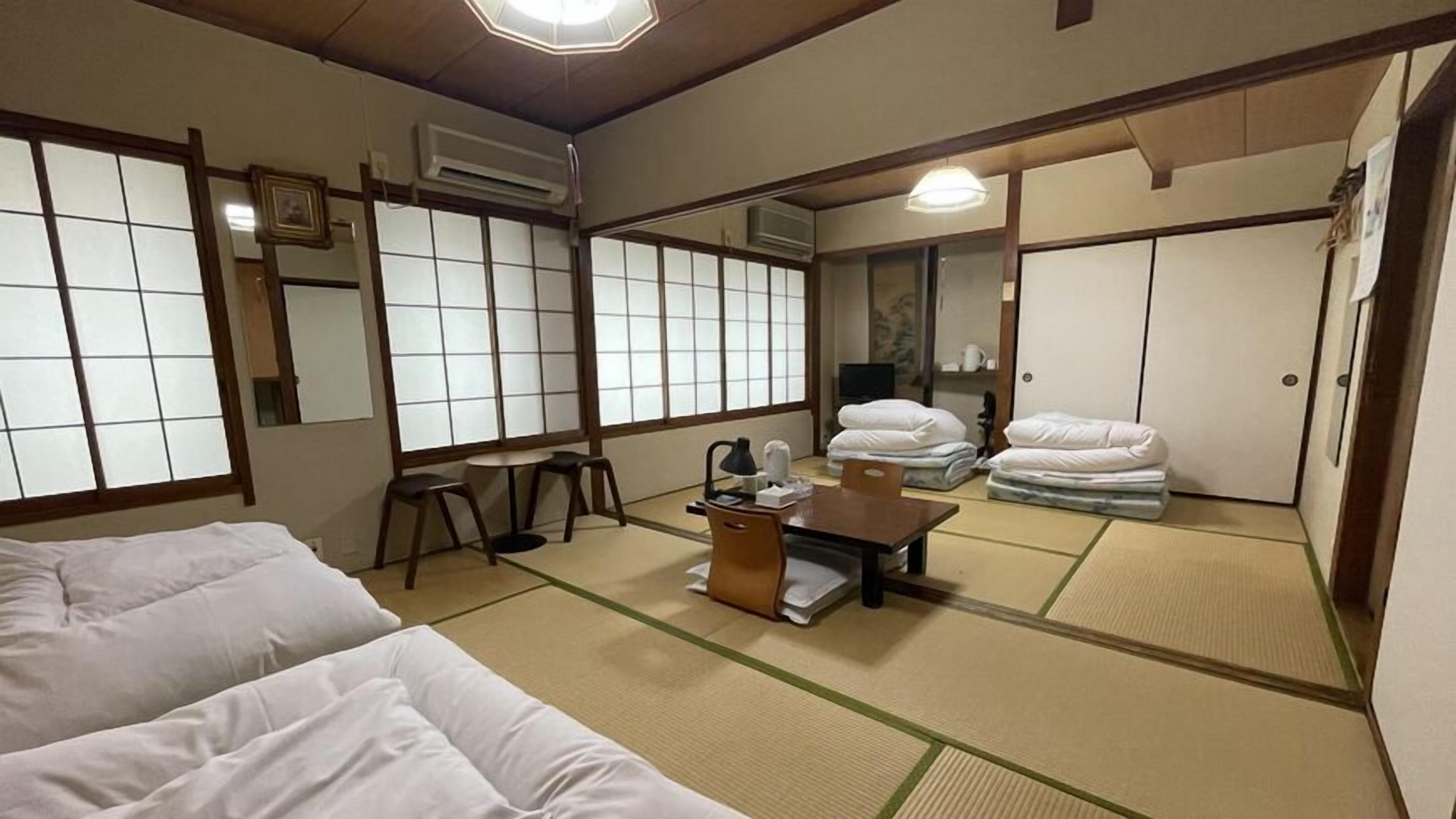 A traditional Japanese room with tatami flooring, a futon bed, a small table with a lamp, and a sliding door.