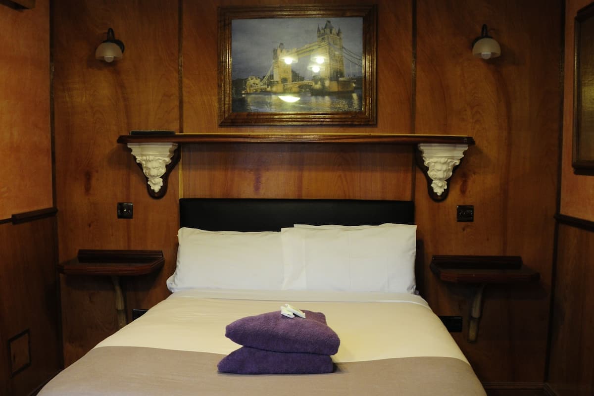Une chambre d’hôtel avec une tête de lit en bois, un tableau représentant un pont et une serviette violette sur le lit.