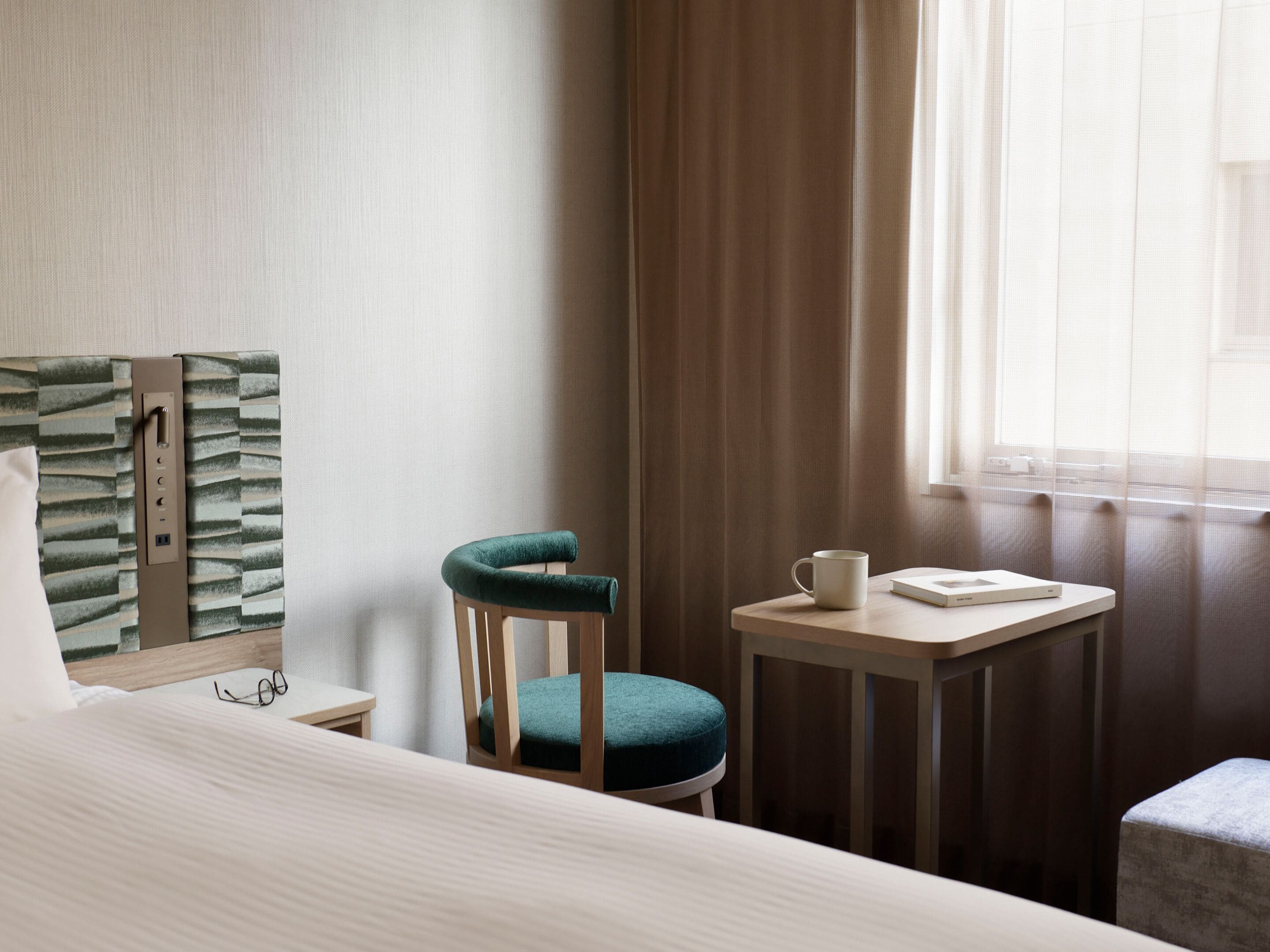 A hotel room with a bed, a chair, a small table, a window with curtains, and a wall-mounted control panel.