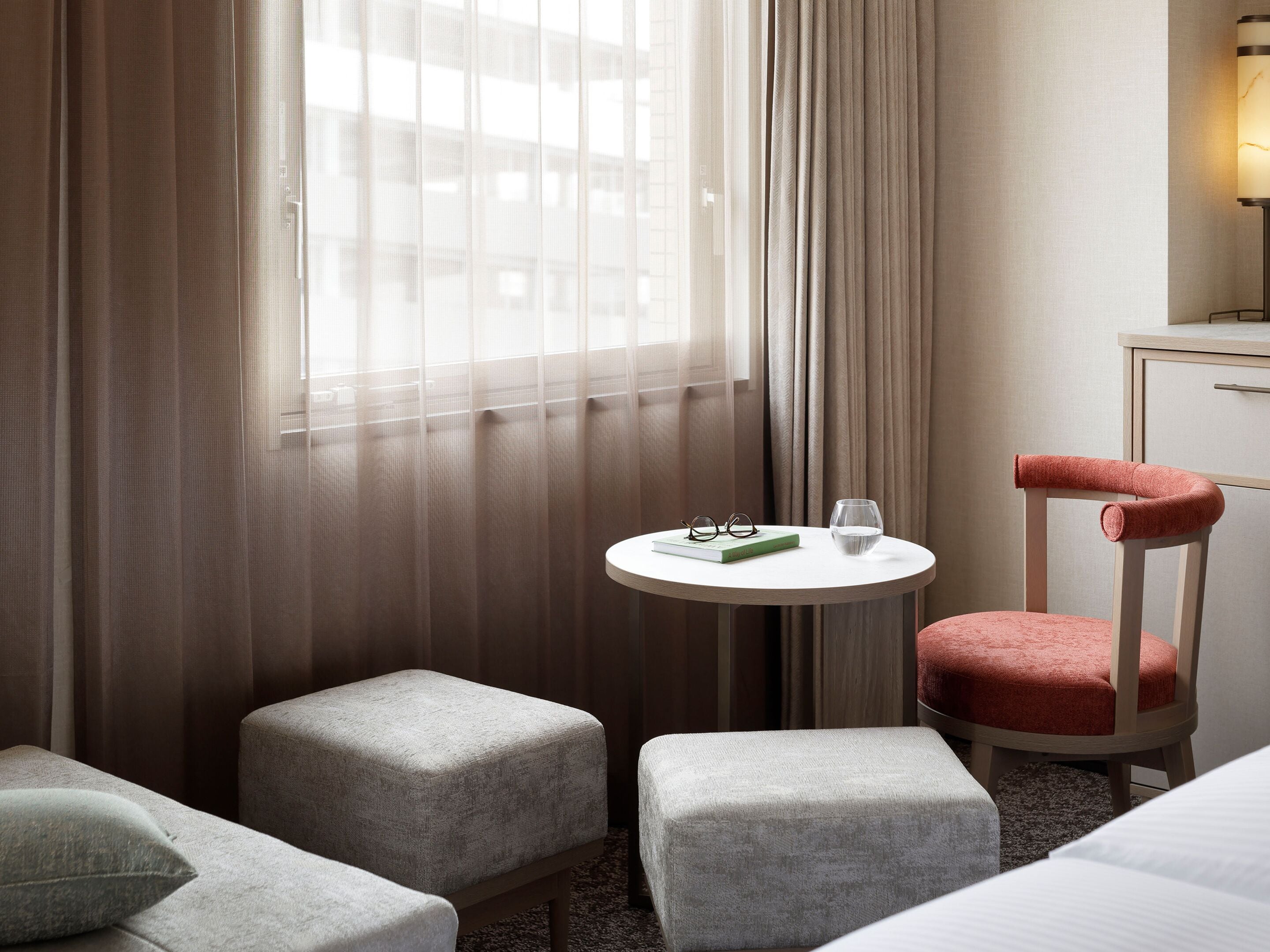 A hotel room with a round table, two ottomans, a chair, and a bed.