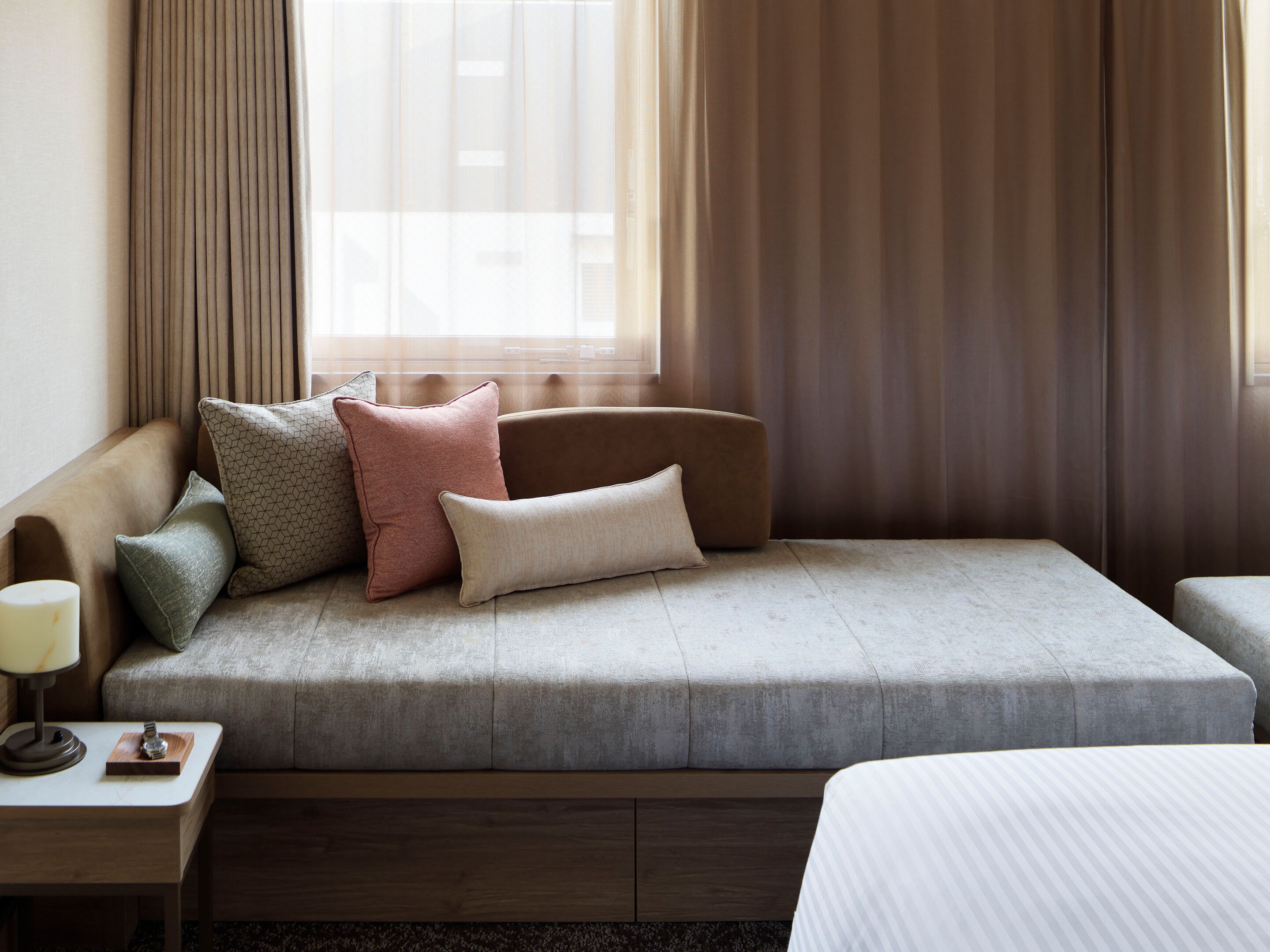 A hotel room with a sofa, pillows, a bedside table with a lamp, and a window with curtains.