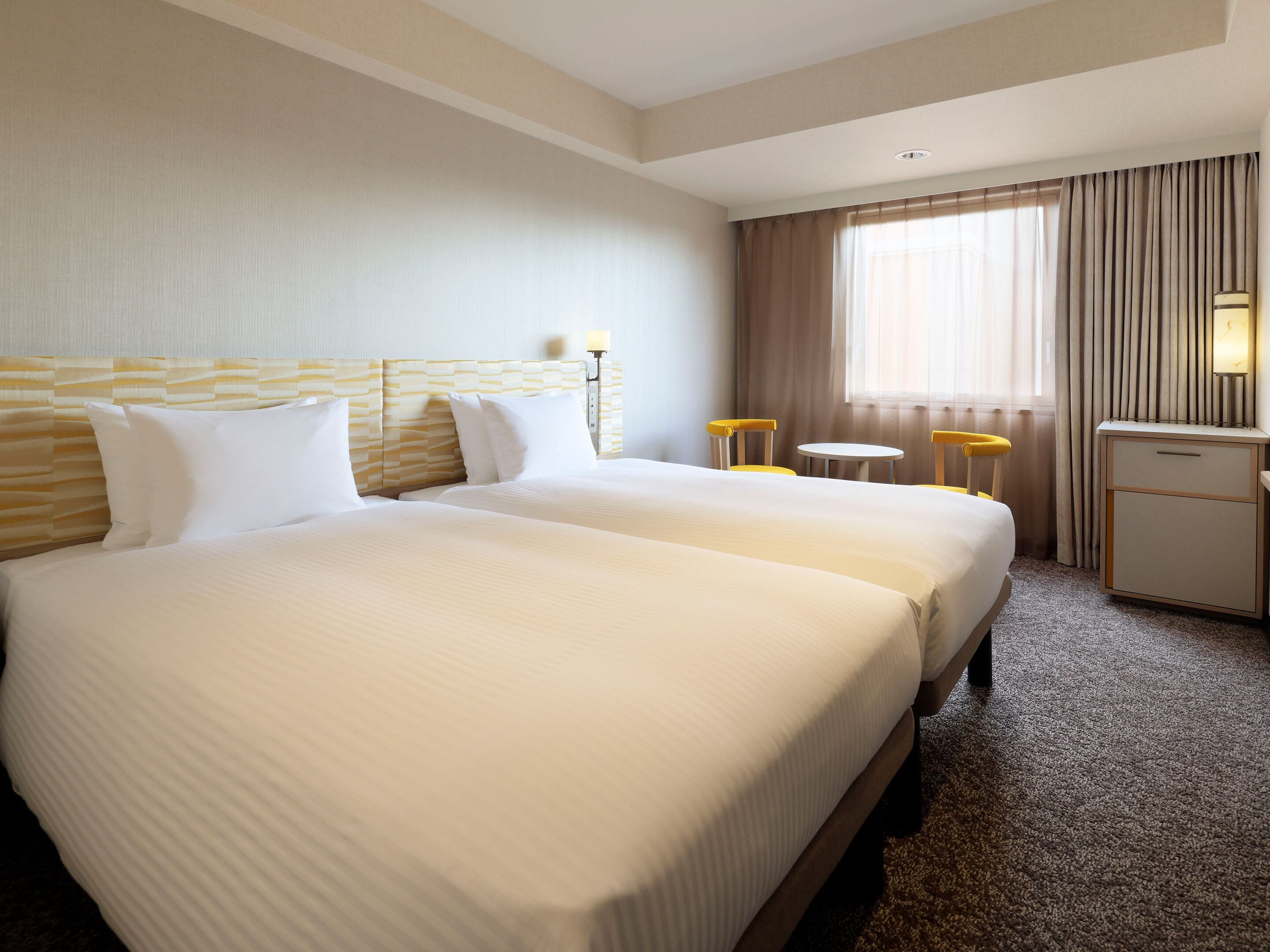 A hotel room with two beds, a small table, a chair, a desk, and a window with curtains.