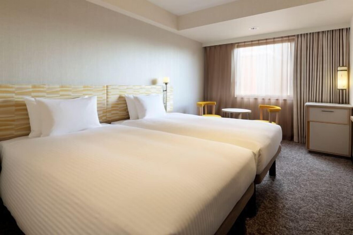 Habitación de hotel con dos camas, un escritorio, una silla y un ventanal con cortinas.