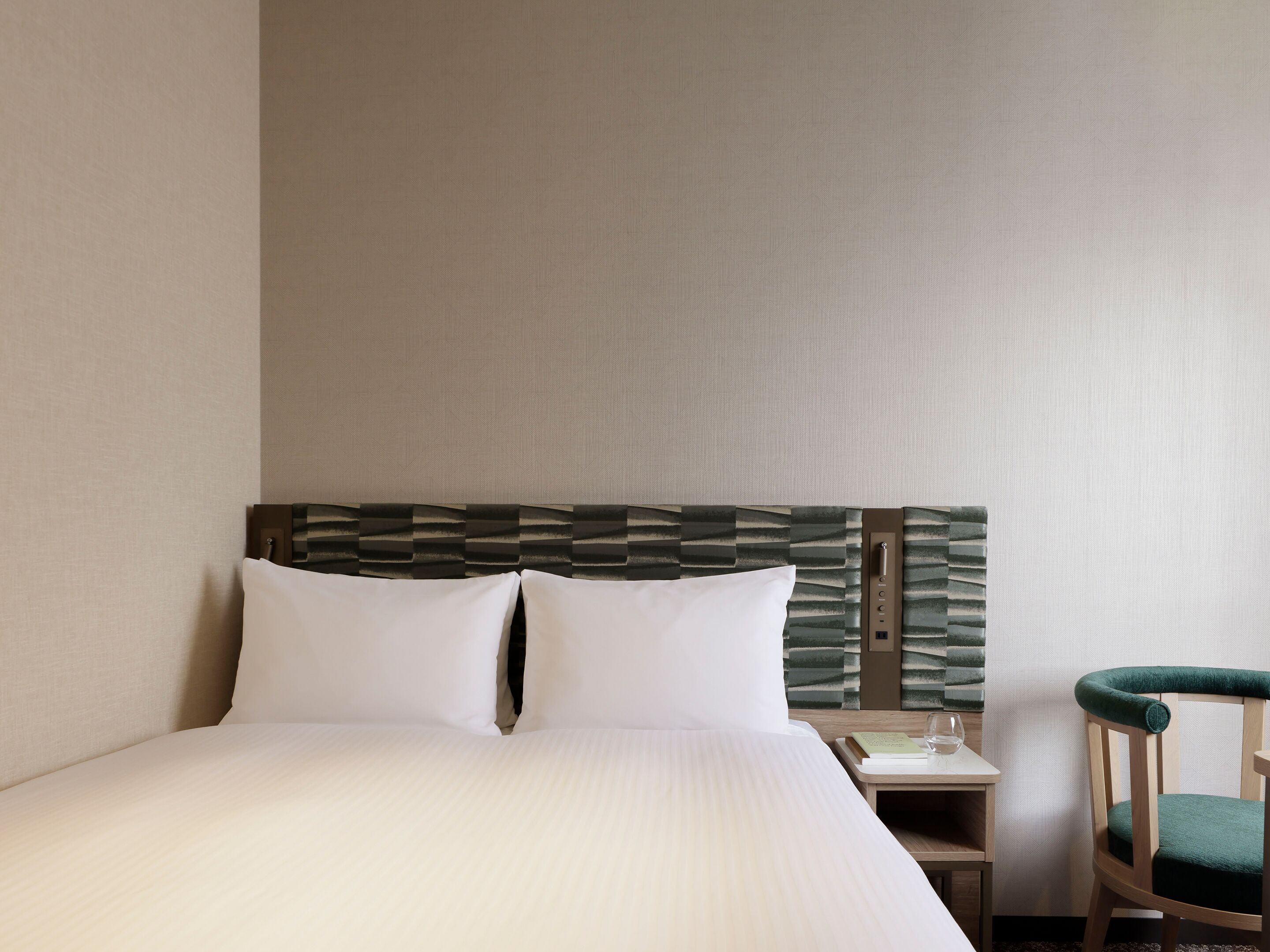 A hotel room with a bed, a nightstand, a chair, and a wall with a textured pattern.