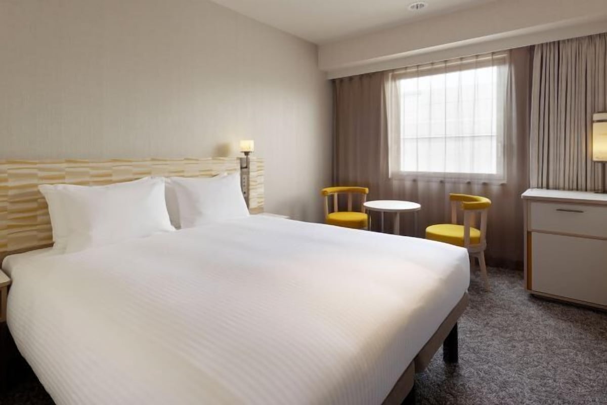 A hotel room with a large bed, a desk, two chairs, and a window with curtains.