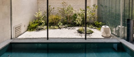 Piscina interior moderna con vista despejada a un jardín ajardinado, una escultura geométrica y una pared de vidrio que refleja el entorno.